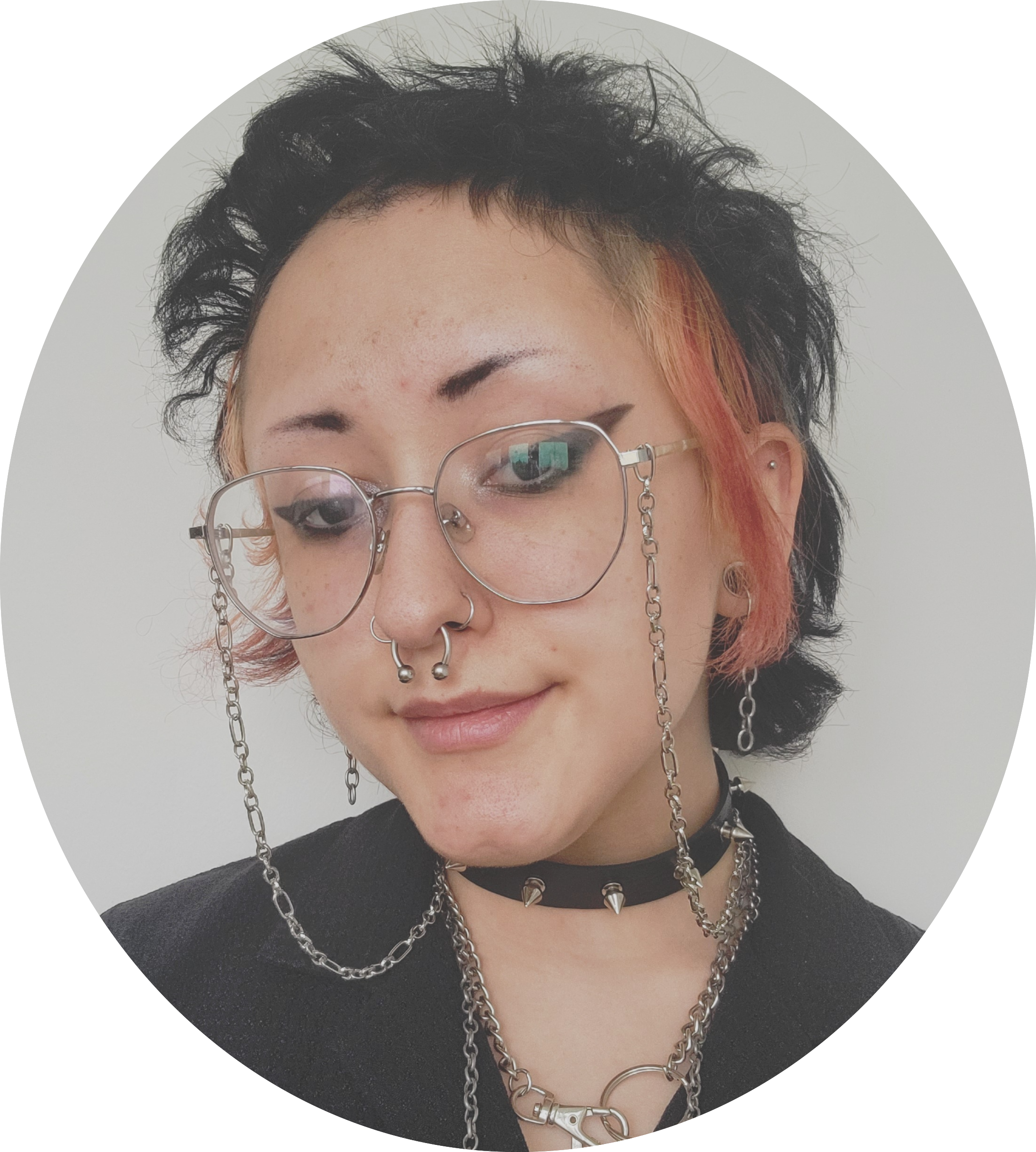 Kaymiss is an androgynous presenting person with winged eye makeup, a nose ring and septum piercing, gauged ears, and short-cropped, fluffy black and pink hair. They are wearing wire rimmed glasses with a chain, a spiked dog collar, a clipped chain necklace and a black shirt.